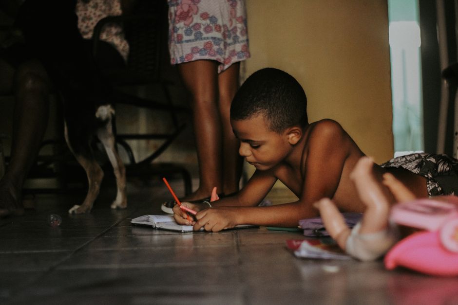 Young boy drawing with red pencil on the floor surrounded by toys indoors.