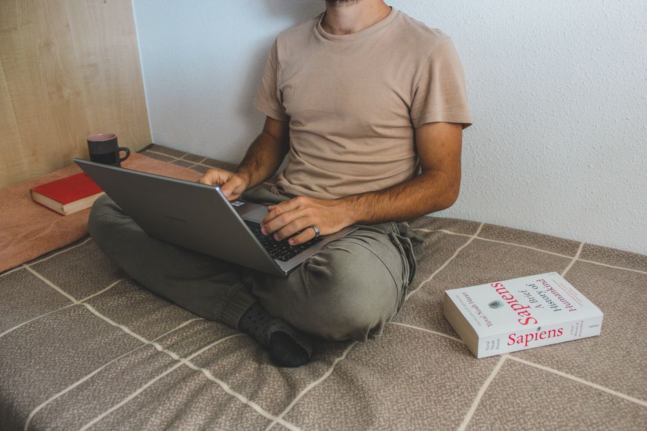Man working remotely on a laptop, surrounded by books in a cozy indoor setting, reflects modern digital lifestyle.