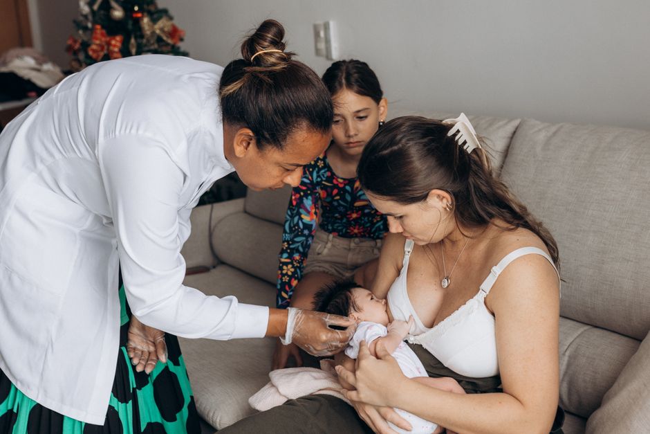 A pediatrician checks a newborn baby held by a mother on a sofa, with a child watching.