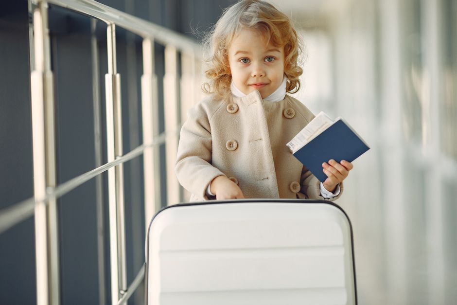Adorable little girl with blond curly hair in stylish beige coat standing with white suitcase and passport in airport corridor